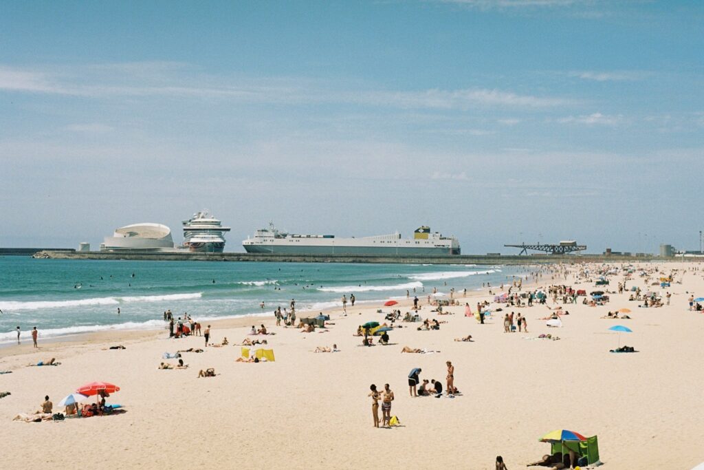 People relax on a sunny beach with ships in the distance.